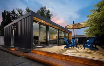 Dunkles Tiny-Gartenhaus mit Holzdeck, Sonnenschirm und blauen Stühlen