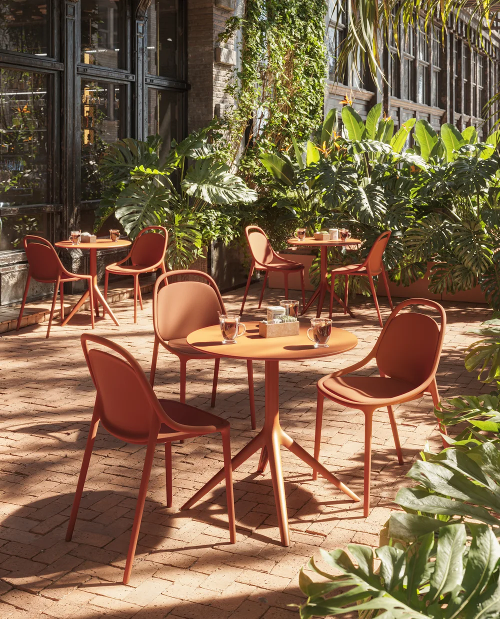 VONDOM B2B: orangefarbene Stühle um runde Tische auf einer begrünten Ziegelterrasse