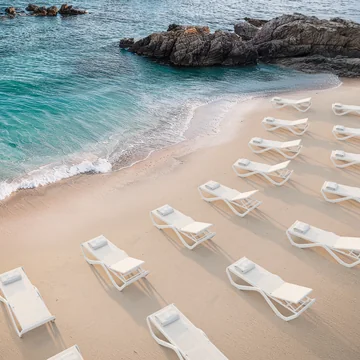 VONDOM LOVE: mehrere weiße Sonnenliegen auf hellem Sand an einer Felsküste