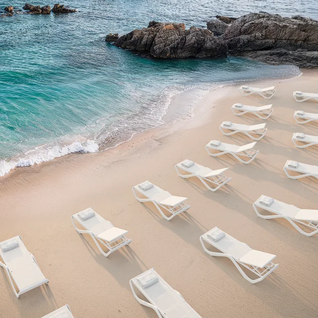 VONDOM LOVE: mehrere weiße Sonnenliegen auf hellem Sand an einer Felsküste